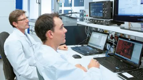 two people with white shirts sitting in from of laptops in a control room