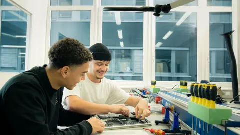 Two people perform technical work on an open laptop at an IT workstation.